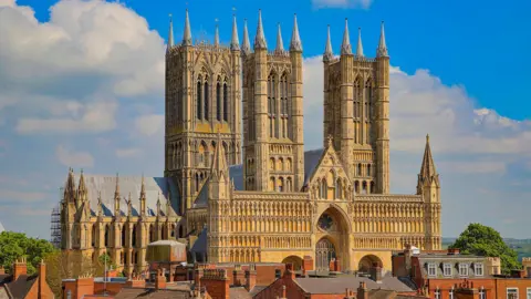 An image of Lincoln Cathedral on a sunny day. The sandstone building is set against a blue sky and surrounded by rooftops of neighbouring buildings.