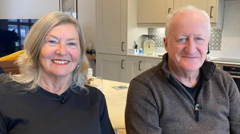 Jacolyn Priestley-Jayes and her husband Tim Jayes sit side by side in their modern kitchen. They are in their sixties and she is wearing a black, long-sleeved t-shirt and he is wearing a dark marl grey half-zip fleece. She has ash blonde shoulder-length hair, he has grey hair which is short and receding.
