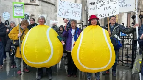 Two women dressed as tennis balls protest outside Royal Courts of Justice with other members of Save Wimbledon Park. They hold various signs with writing such as "Land In Trust For All Of Us!".