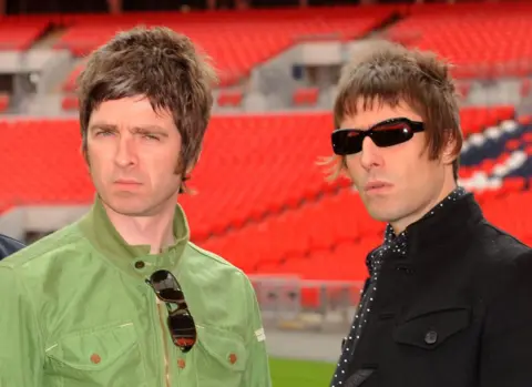 PA Noel and Liam Gallagher frown at the camera in a photo taken in 2008. Noel is wearing a green jacket and Liam is wearing a black one. He wears sunglasses. They both have modern hairstyles.