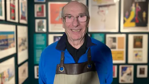 BBC Ian is standing in the museum with framed pictures on the walls behind him. He is wearing a blue jumper and is smiling at the camera.