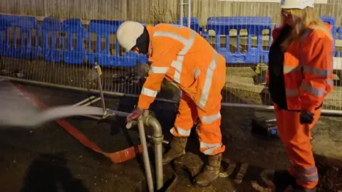 Two workers in orange Thames Water uniforms work on a grey pipe losing water