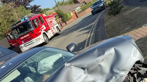 Shropshire Fire and Rescue Service Half of a silver car is pictured up close, the bonnet has been crushed and the windscreen is smashed. The driver air bag has also been deployed. A fire engine is parked behind the car, as well as another vehicle, a black Audi. All of the vehicles are on a residential street. 