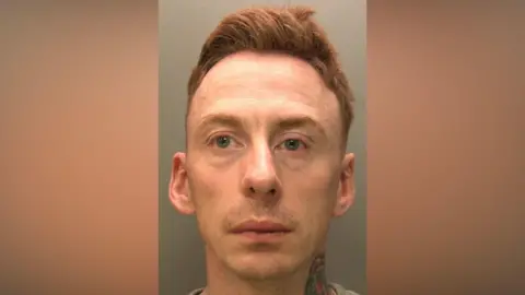 Mugshot of Brian Smith. He has ginger hair which is gelled up and is looking sad and at the camera. He has a neck tattoo of a red rose. He is clean shaven and is wearing a grey jumper. 