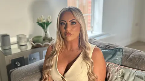 BBC A woman with long blonde hair and blue eyes is looking directly into the camera, unsmiling. She is wearing a light-coloured sleeveless v-neck top. The background is out of focus but she is sitting on a grey sofa with light green cushions, behind her is a table with white flowers on it and a window. 