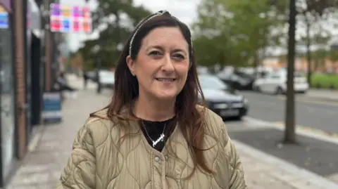 Amanda Bower has long brown hair and is wearing a hairband, gold coloured jacket and black top. She is also wearing a silver necklace that says Amanda.