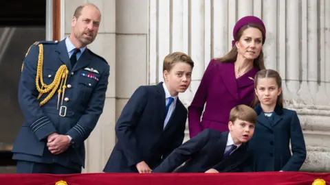 William, Prince of Wales looks on while Catherine, Princess of Wales and their three children lean to the right as they stand on the balcony of Buckingham Palace and gaze into the distance to see better.