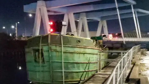 Close-up shot of a barge wedged under a bridge. The barge has a green hull and mooring ropes dangling down. There are railings to the right side of the image.