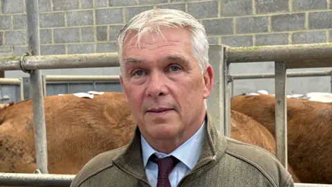 A man with grey hair, wearing a blue shirt, dark tie, and khaki quarter-zip top standing in front of a metal fence with cows behind him.