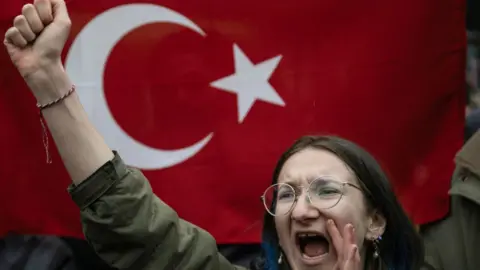 A woman shouts and holds her fist in the air - a Turkish flag is behind her