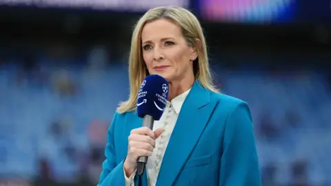 PA Media Gabby Logan pitch side ahead of a football match, holding a microphone and wearing a blue jacket and white top.