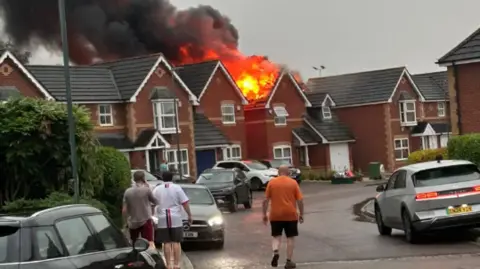 Onlookers watch as the roof of a house burns fiercely.