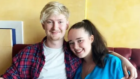 A young couple, he has blond hair, a tartan shirt and a white T-shirt underneath. She is in a light blue top and has long dark hair. They are sitting on a reddish sofa.