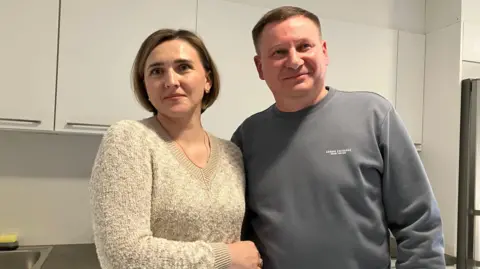 Dariia and Ihor Zhurba stand in a communal kitchen at a nuclear power plant in Kyiv, facing the camera. Dariia is wearing a beige V-neck jumper, while Ihor is wearing a grey sweatshirt. 