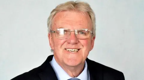 Shropshire Council A smiling man with grey hair and silver rimmed glasses in a dark suit with a white tie and a light grey background