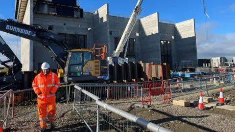 Peterhead building site, where the connector for the subsea cable is being built - Construction worker in high‑visibility orange gear walks across a large building site with heavy machinery, barriers, and a partially built concrete structure in the background.