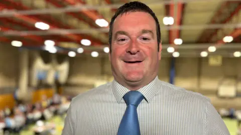 Head and shoulders image of Ryan Coogan. He has short brown hair and is wearing a grey and white striped shirt and blue tie.