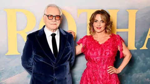 Getty Images Cox and his wife Nicole Ansari Cox at the premier of Glenrothan - Brian is in a navy velvet blazer and Nicole wears a vibrant red dress.