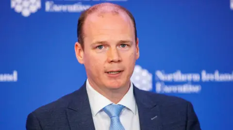 Gordon Lyons is standing in front of a blue background. There is white writing that reads "Northern Ireland Executive". 