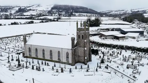 People make their way through snow to Mass at Holy Cross Catholic Church in Killeshin, County Laois, Ireland., drone shot of church and surrounding fields and hills covered in snow