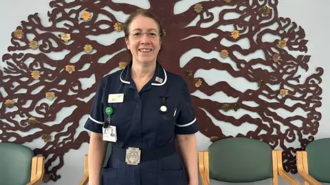 Mel Taylor, palliative care specialist, is wearing her navy nurses uniform. She has brown hair tied back and wears glasses. Behind her on the wall is the silhouette of a brown tree with shining gold leaves on it.