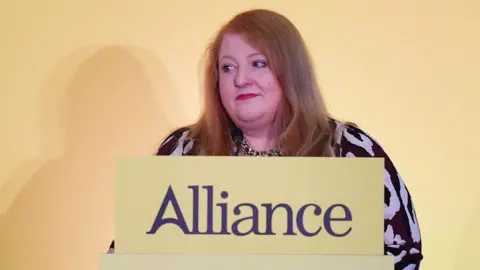 Naomi Long, a woman with shoulder-length ginger hair and red lipstick, is standing in front of a yellow podium which is topped with a sign showing the Alliance Party logo. She is wearing a black and taupe patterned dress and a chunky necklace.