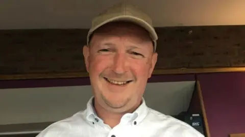 Glenn Quinn wearing a cream coloured cap and a white shirt. He is smiling at the camera. 