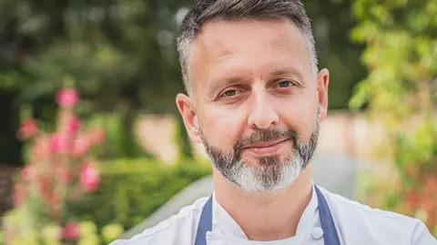 Head and shoulders image of Mark Birchall in his chef whites with a navy blue apron in the restaurant's garden. He has short dark hair, greying at the sides and  beard