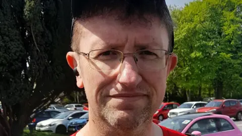 Tom Wright has short reddish hair and is wearing a baseball cap. He has wire-rimmed glasses and is standing in a car park