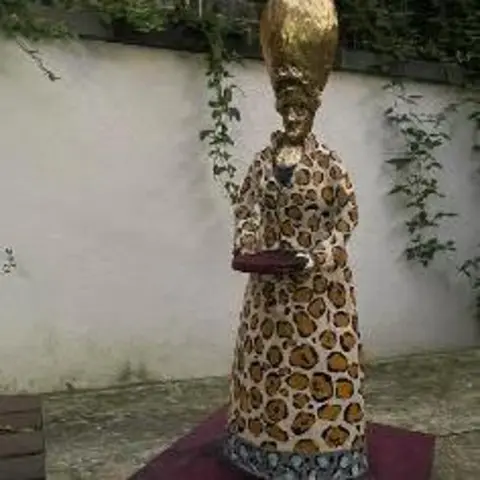 Staffordshire Moorlands District Council A gold-coloured statue of the goddess Ninkasi, on a purple coloured plinth. The statue is stood in a courtyard, with a white-painted wall behind which has some vegetation hanging down it.