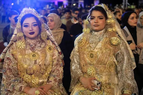 ABDULLAH DOMA / AFP / GETTY IMAGES Two women are seated wearing matching golden-coloured dresses and headdresses.