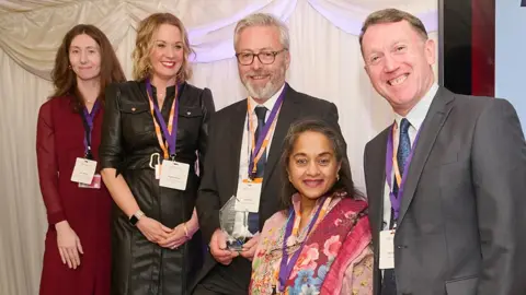 LCC Lancashire County Council Trading Standards food team at the awards ceremony. Three women and two men, all dressed smartly, are wearing purple lanyards and smiling at the camera