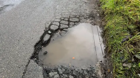 A large water-filled pothole on a road and next to a grass verge. There are cracked bits of tarmac all around it.