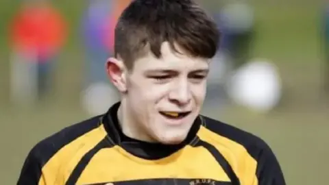 Aaron Bell has brown hair and wears a black and yellow rugby jersey and gum shield