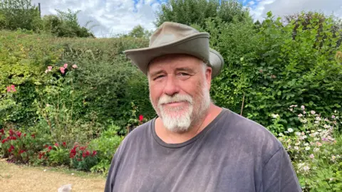 Tim Hoverd wearing a grey hat and dark grey t-shirt is stood in a garden. He has a large hedge with a flower bed behind him. He also has a white beard and is smiling at the camera.