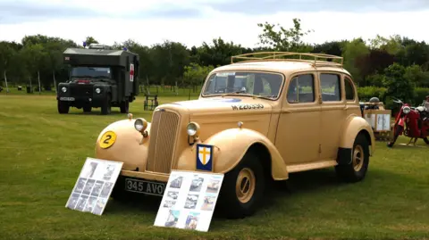 MVT A yellow/gold car with a long bonnett and wheel arches. It is displayed in a field alongside other vehicles. 