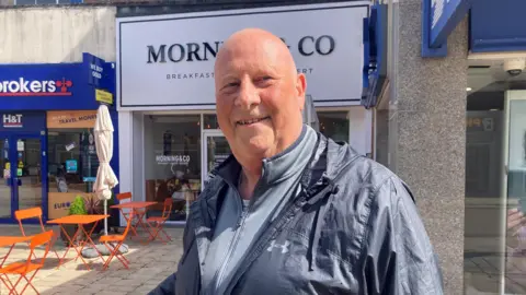 A bald man is smiling. He is wearing a grey zip-up sweater and waterproof over it. There are orange tables and chairs behind him.