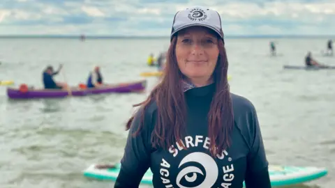 STEPHEN HUNTLEY/BBC A woman wearing a “Surfers Against Sewage” wetsuit and hat stands in shallow seawater, with paddleboarders and a canoe visible on the water in the background.