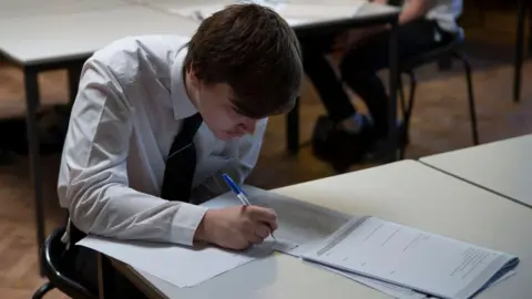 Getty Images A boy in a white shirt and school tie is sat at a school desk writing on a sheet of paper with a blue biro, with a booklet open in front of him.