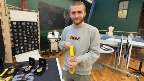 A man with a shaved head and a beard, with a grey jumper, smiles as he holds a bronze medal, He is standing in a hall next to a table with other medals on display.
