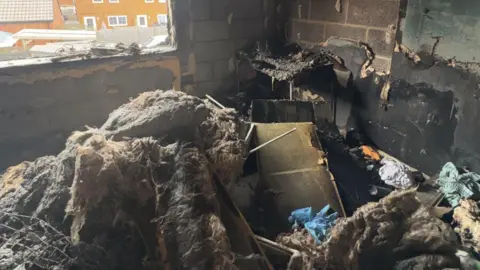 Heavily burned debris, furniture and insulation piled up on the floor of a bedroom. The window has shattered and all the paint and plaster have been stripped from the walls, leaving only blackened breeze blocks. 