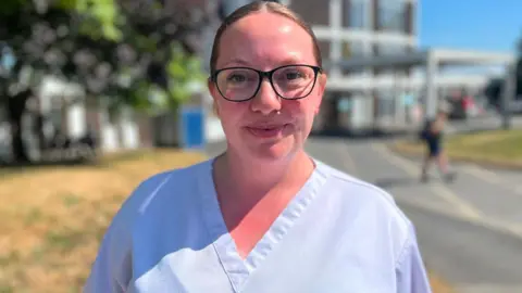 The picture shows a person standing outdoors in front of a modern hospital-style building, wearing light-coloured medical scrubs and glasses, with the photograph focused sharply on the person while the background is blurred. The scene is brightly lit by sunshine and includes a paved walkway, dry grass, trees and part of the hospital complex, with another figure visible in the distance near the building.