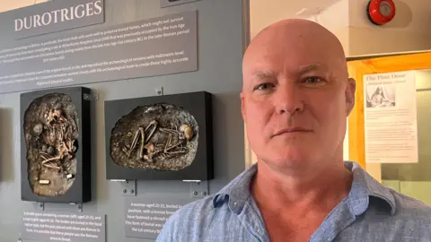Dr Martin Smith stands in front of a display board showing 3D scale replicas of skeletons