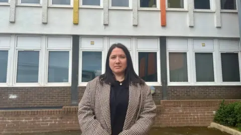 A woman with dark hard and a chequered coat stands in front of the block of flats