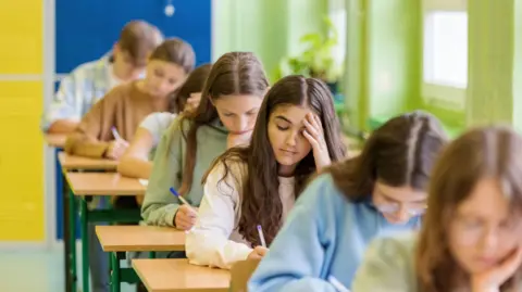 Six people taking an exam in an exam hall. One girl is in focus putting her hand on her head. She is writing an answer.