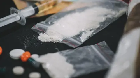 A clear plastic bag of white powder with some spilling on to the black table top. There is also a syringe and several white and orange pills on the table. 