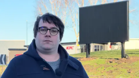 A man wearing black rimmed glasses and a dark blue jacket with a grey jumper stands in front of a roundabout where a 17ft high LED display has been installed. A branch of KFC can be seen in the distance.