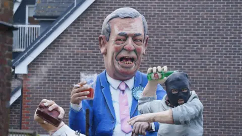 PA Media A large model of Nigel Farage holding a cigarette and a pint of beer. He is wearing a blue suit and pint tie. Next to him is a model of a person in a grey jumper wearing a black hood, throwing an empty glass bottle of Stella Artois