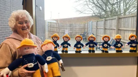 Maureen Atkinson holding two large knitted firefighter toys. She has white hair and is wearing a fleece.  The knitted firefighters have yellow helmets and blue coats. A line of seven smaller toys are standing on the windowsill inside the fire station.