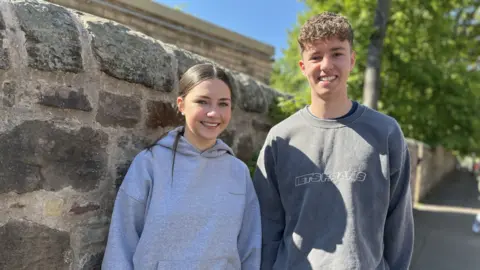A girl and a boy in grey sweatshirts stand against a stone wall